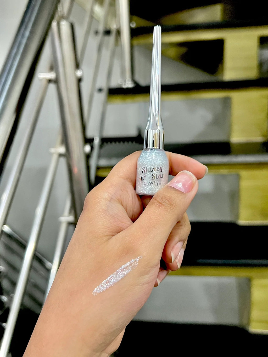 Hand holding a silver and white cosmetic product with visible brand name, against a blurred indoor background.