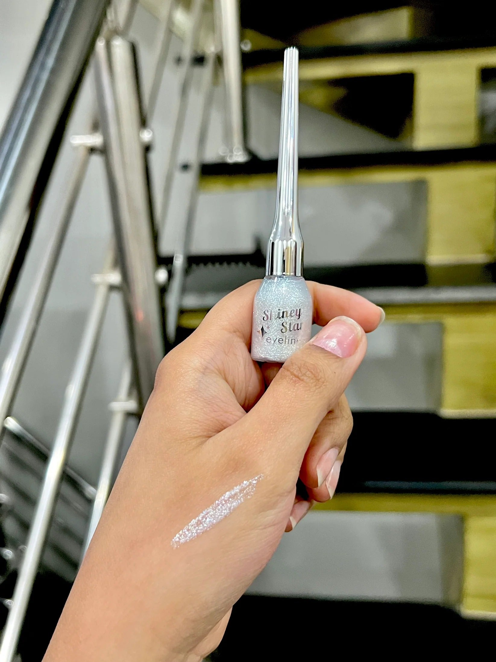 Hand holding a silver and white cosmetic product with visible brand name, against a blurred indoor background.