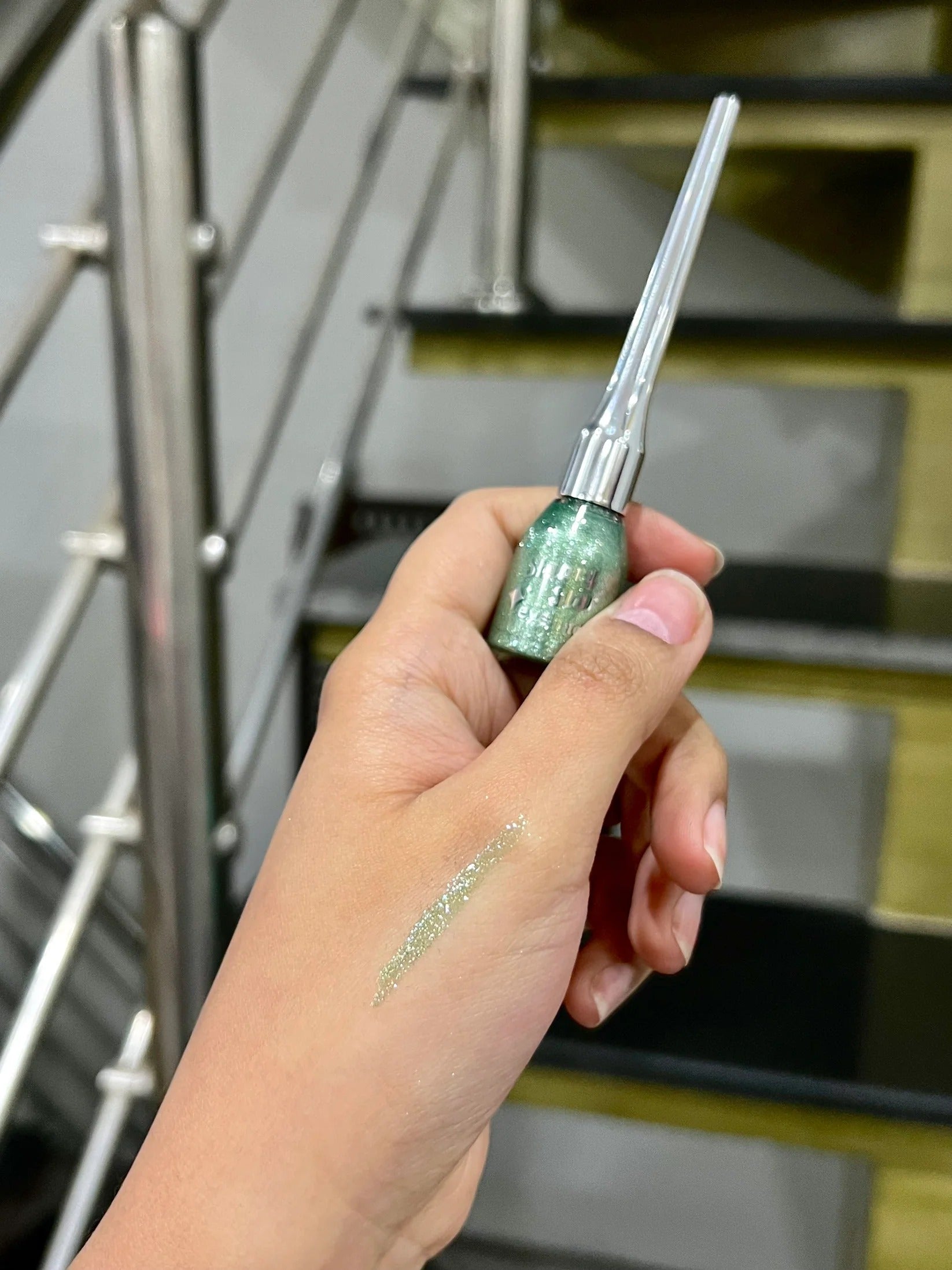 Hand holding a green and silver eyeliner pen with a blurred staircase background