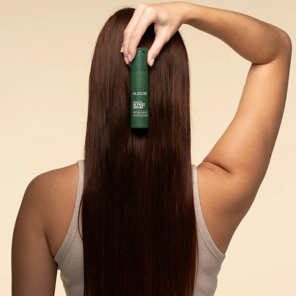 Woman holding a green bottle of Blesso hair product against her long, straight hair on a beige background