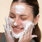 A close-up of a woman with white facial cleanser or shaving cream applied to her face and hands, gently washing her face against a plain beige background.