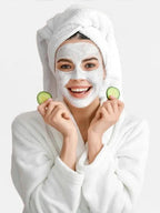 Woman with a facial mask holding cucumbers, wrapped in a towel, on a white background