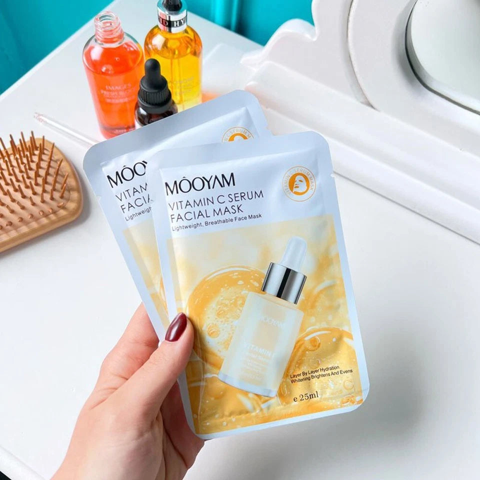 Two Mooyam Vitamin C Serum Facial Mask packages held by a hand on a white surface with skincare products.