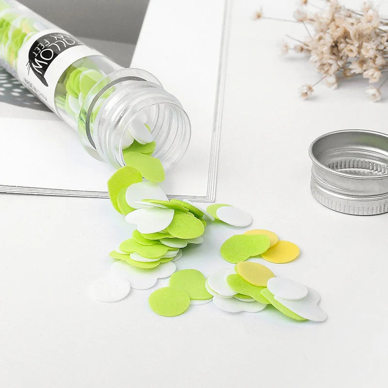 Green and white circular confetti spilling out of a transparent container on a light surface.